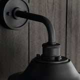Close-up of the Caldton Outdoor Wall Light in matt black, showing the curved arm, knurled fitting, and shade top against a wood-panelled wall.