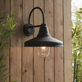Endon Brackworth black outdoor wall light with gooseneck arm, textured metal shade and clear glass tube around an E27 filament bulb, on a wooden fence.
