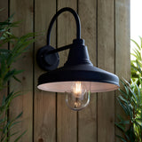 Endon Brackworth outdoor wall light in textured black with a gooseneck arm, barn-style shade and clear glass globe housing a filament bulb, on a wooden fence.