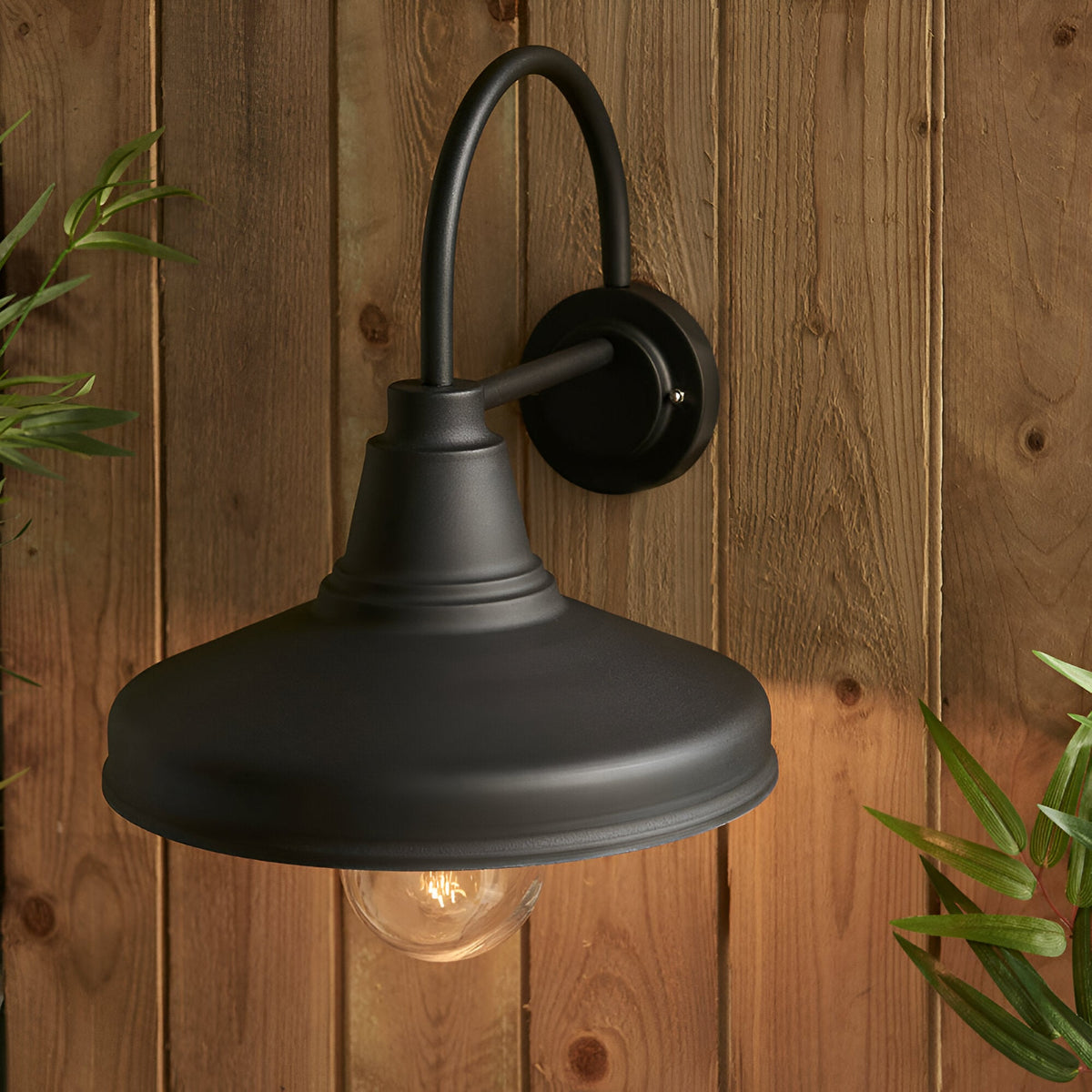Endon Brackworth outdoor wall light in textured black metal with a gooseneck arm and barn-style shade, mounted on a wooden plank wall beside foliage.