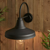 Endon Brackworth outdoor wall light in textured black metal with a gooseneck arm and barn-style shade, mounted on a wooden plank wall beside foliage.