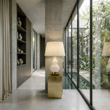 A modern interior showcases the Lumetra 1 Light Sculpted Alabaster Table Lamp in Brass & Soft White on a gold pedestal by tall windows with views of greenery and a pool, while built-in shelves on the left display decorative vases.