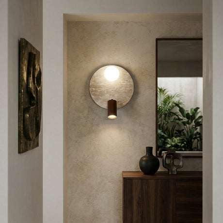 The Korvane 2 Light Travertine Wall Light in walnut brown and light beige shines above a wooden console with vases and a sculptural piece, while a mirror behind reflects lush green plants.