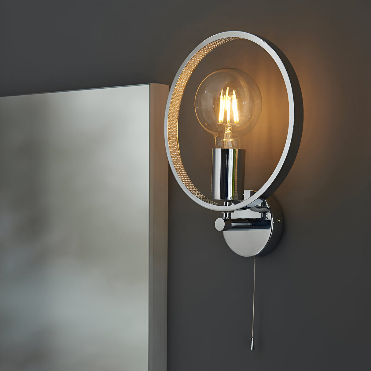 Endon Carlcombe chrome bathroom wall light with faceted circular ring, E27 globe bulb and pull cord switch, mounted beside a mirror on a grey wall.