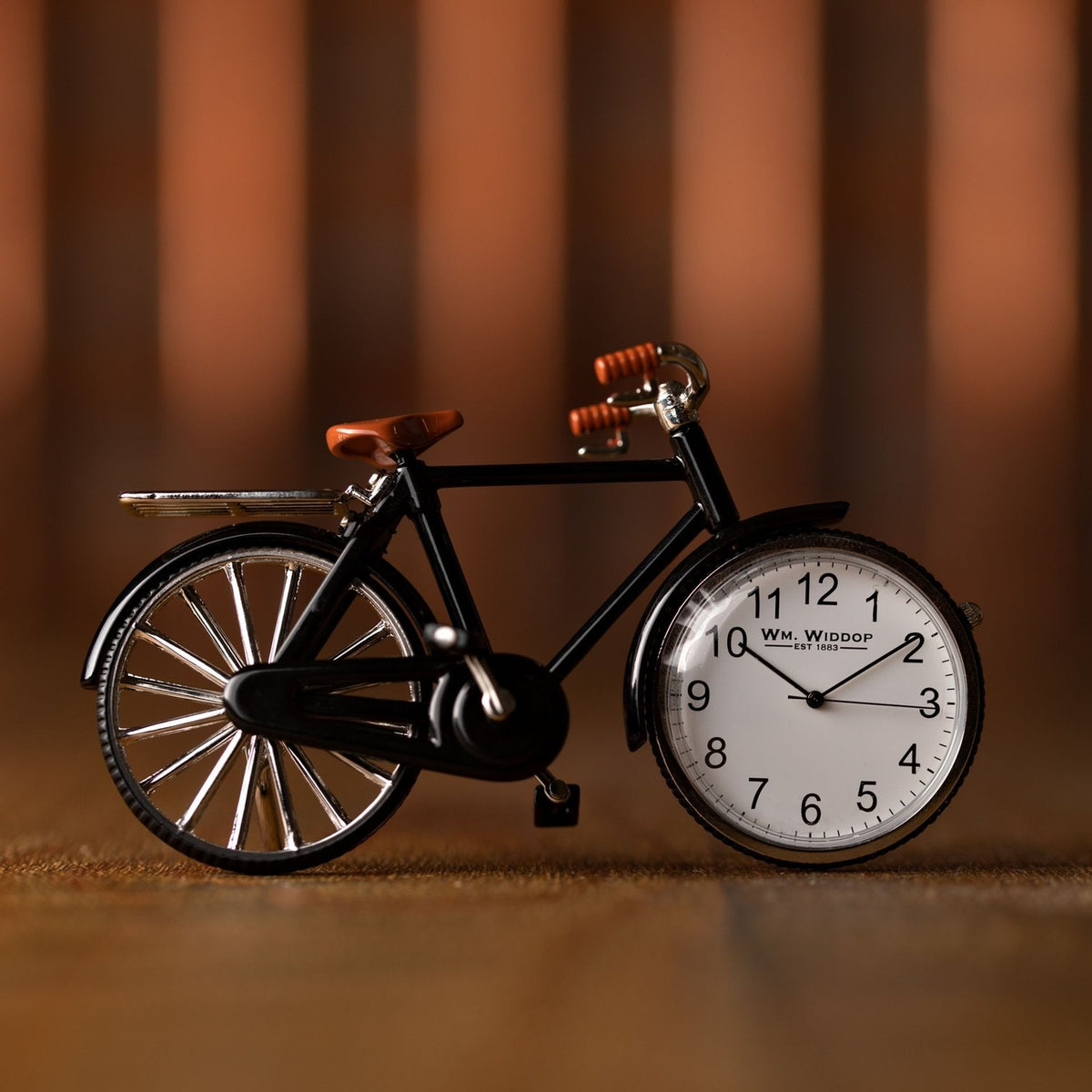 Miniature mantel clock shaped like a black pedal bike, with silver-toned spokes, rear rack and handlebars, a brown saddle and grips, and the white Arabic numeral dial set into the front wheel.