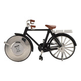 Black miniature pedal bike mantel clock with silver spokes, handlebars and pedal cranks, brown saddle and grips, clock face set into the front wheel, on a white background.