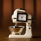 Miniature mantel clock shaped like a food mixer, beige glossy metal body with silver mixing bowl, beaters, and a white analogue clock face with black Arabic numerals set into the mixer head.
