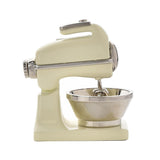 Miniature food mixer mantel clock in glossy beige metal with a silver mixing bowl, beaters, and chrome detailing, shown side-on against a plain white background.