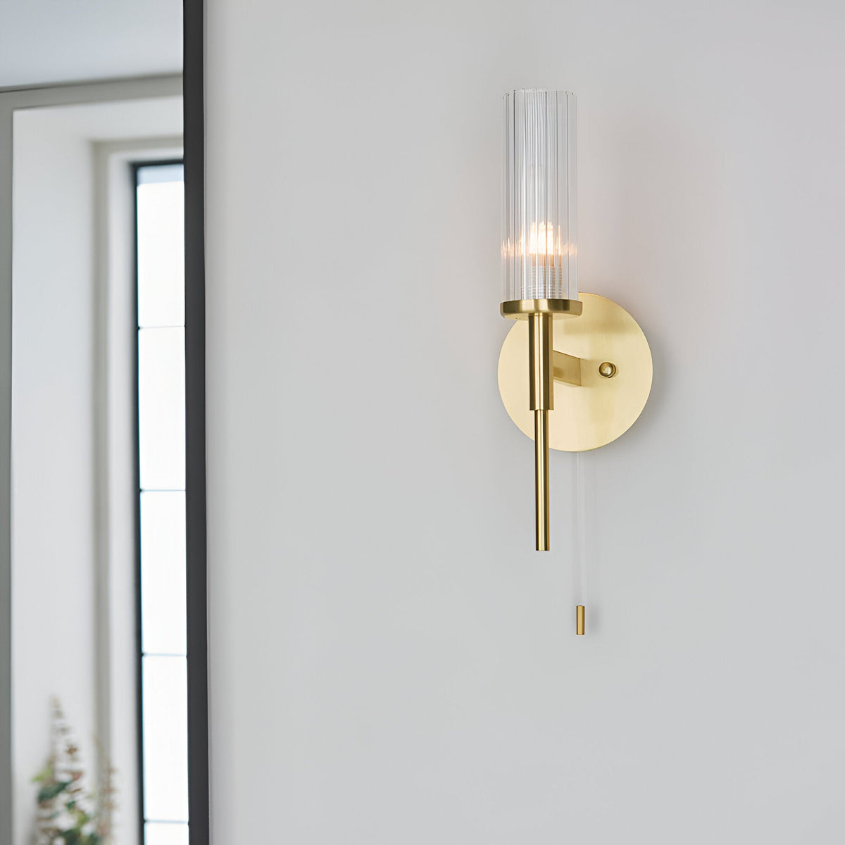 Endon Wren satin brass bathroom wall light with clear ribbed cylindrical glass shade and pull switch, mounted on a white wall beside a mirror.
