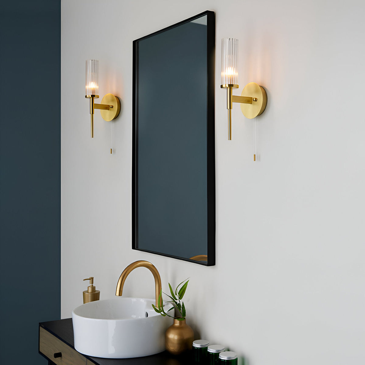 Endon Wren satin brass bathroom wall lights with clear ribbed glass cylinder shades, mounted in a pair beside a black-framed mirror above a brass tap and basin.