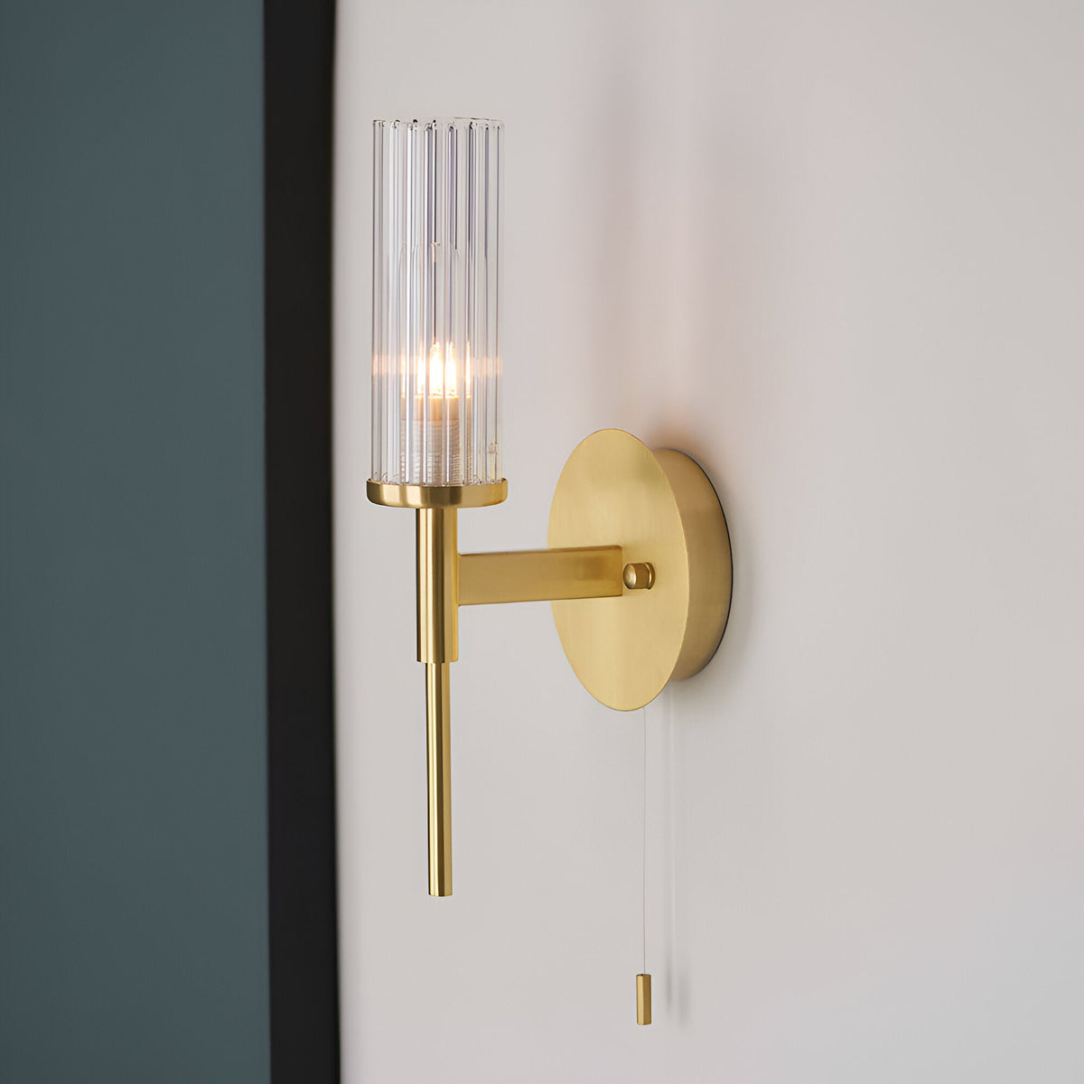 Endon Wren bathroom wall light in satin brass with a clear ribbed cylindrical glass shade and pull switch, mounted beside a dark panel.