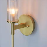 Endon Wren bathroom wall light in satin brass with ribbed clear glass cylinder shade, round wall plate and illuminated G9 bulb, mounted on a light wall.