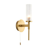 Endon Wren bathroom wall light in satin brass with a clear ribbed cylindrical glass shade, round backplate and pull switch, on a white background.