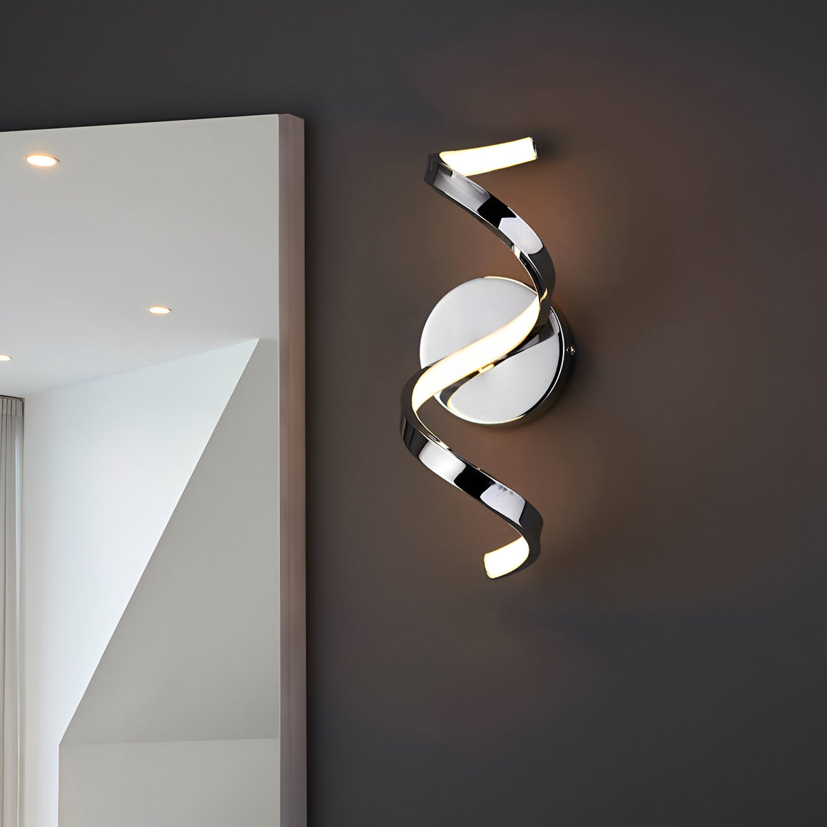 Endon Aldleigh LED wall light in chrome with a tall twisting spiral form and warm white glow, mounted beside a mirror on a dark grey wall.