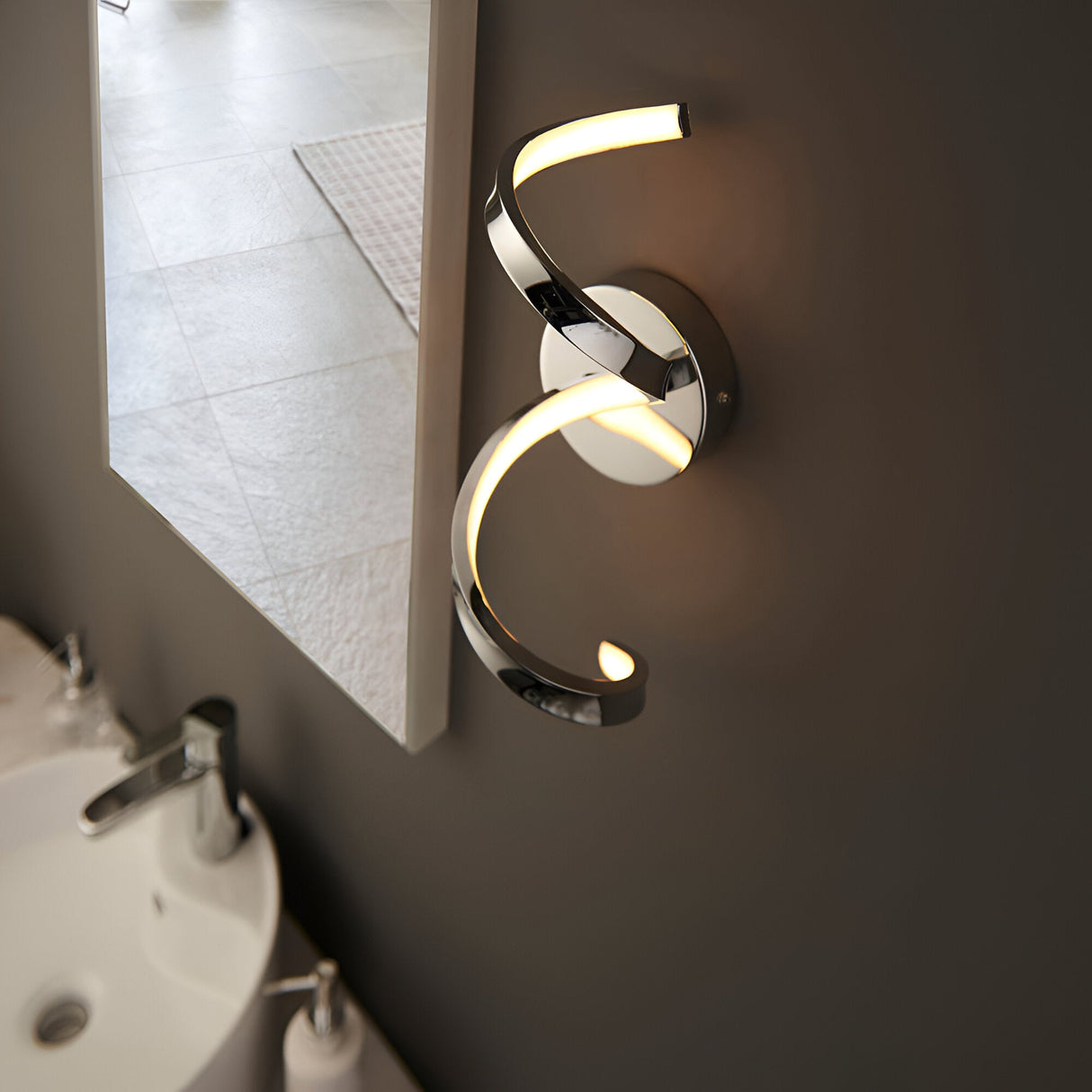 Endon Aldleigh LED wall light in chrome with a twisting spiral form and warm white glow, mounted beside a bathroom mirror above a basin.
