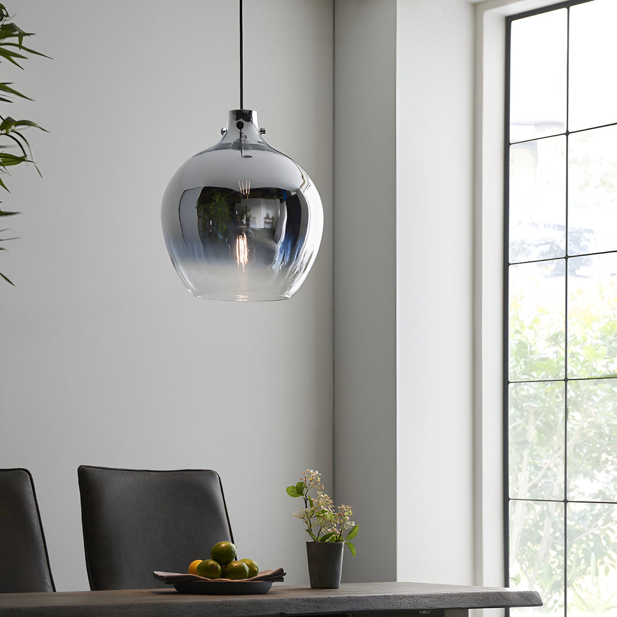 Castworth Glass Pendant in chrome, a single globe-shaped light with ombre finish and visible bulb, hanging above a dining table in a modern room.