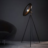 Endon Bramshaw tripod floor lamp in matt black with nickel details and large angled dome shade, beside a dark velvet armchair against a grey wall.