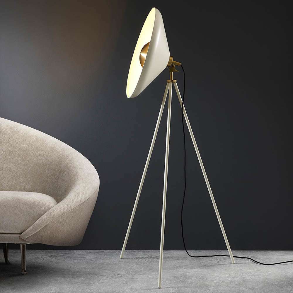 Endon Bramshaw floor lamp in white with brushed brass details, angled conical shade on a tripod stand, beside a cream armchair in a dark room.