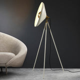 Endon Bramshaw floor lamp in white with brushed brass details, angled conical shade on a tripod stand, beside a cream armchair in a dark room.