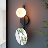 Endon Cliffshaw matt black bathroom wall light with opal glass sphere and adjustable round magnifying mirror, mounted on a dark wall beside foliage.