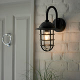 Chartgrove Outdoor Wall Light in black, caged metalwork over a clear glass shade with an E27 bulb, mounted on a whitewashed brick wall by a door.