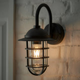 Chartgrove Outdoor Wall Light in black aluminium, with curved arm, dome shade and caged glass shell housing a lit E27 bulb, mounted on a painted brick wall.