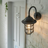 Chartgrove Outdoor Wall Light in black aluminium, showing a caged lantern shade with a curved arm mount on a white brick wall beside a front door.