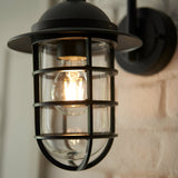 Chartgrove Outdoor Wall Light in black, close-up of caged metalwork over clear glass shade with lit E27 bulb, mounted on brick wall.