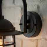 Close-up of the Chartgrove black outdoor wall light showing the aluminium mount, curved arm and caged metal shade over clear glass on a stone wall.