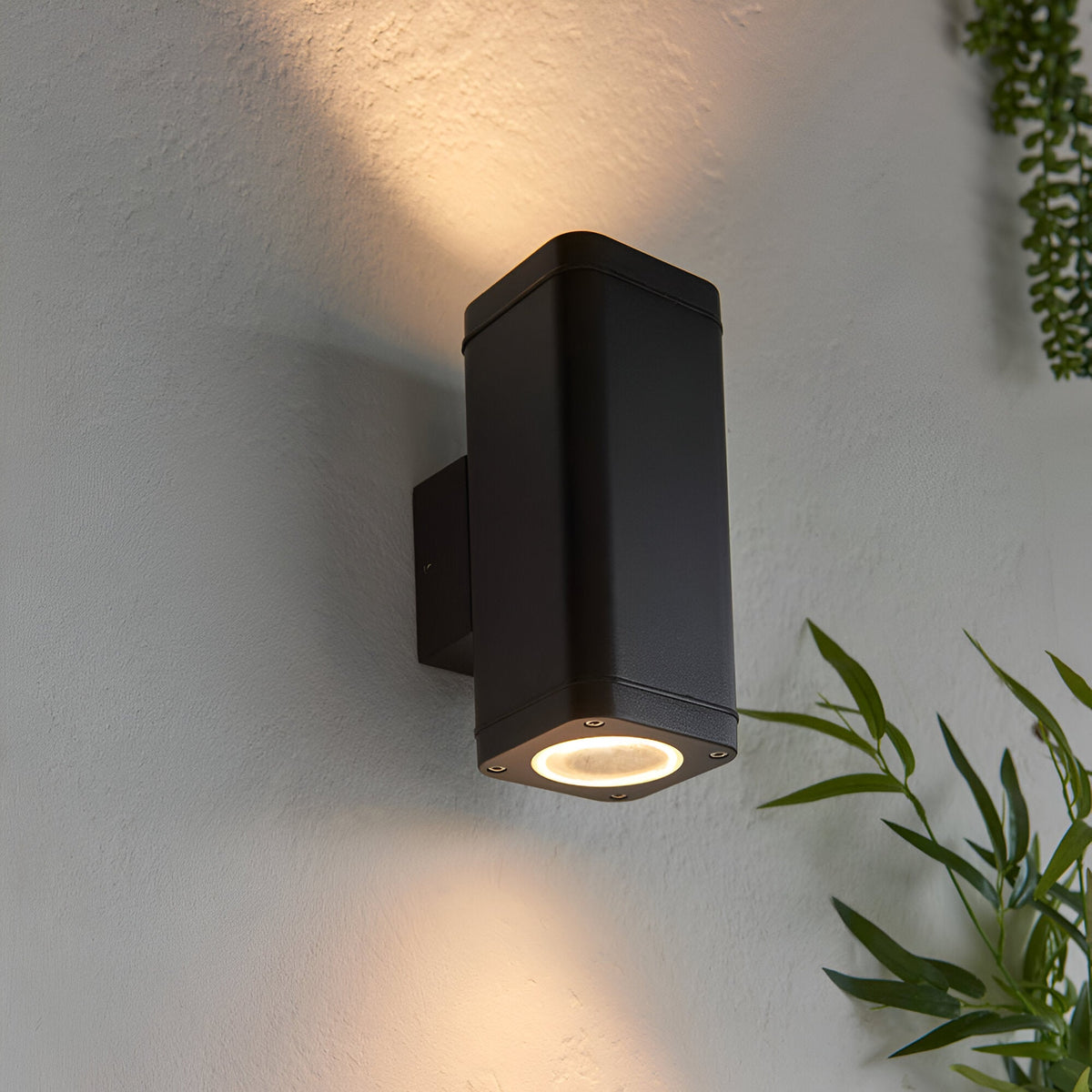 Carlford 2 Light Outdoor Wall Light in black aluminium, rectangular design casting up and down light, mounted on a textured wall beside greenery.
