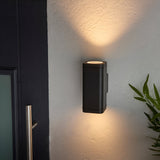 Carlford 2 Light Outdoor Wall Light in black aluminium, mounted beside a front door, casting warm up and down light on a textured wall.