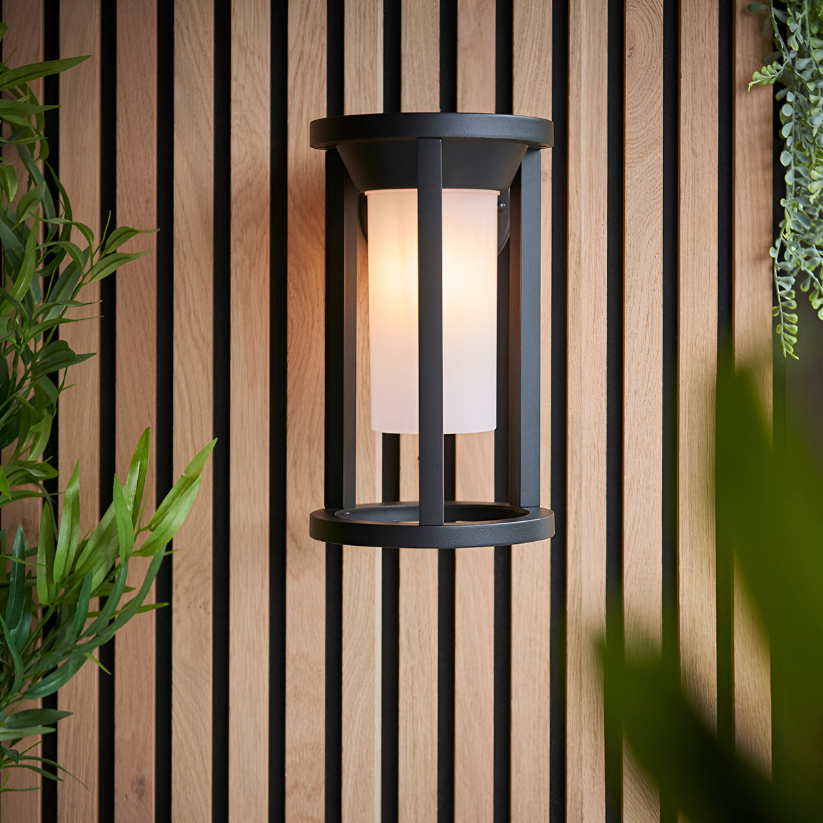Endon Ashdale outdoor wall light with black aluminium frame and white polycarbonate shade, mounted on a wood slat wall beside greenery.
