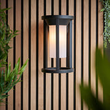 Endon Ashdale outdoor wall light with black aluminium frame and white polycarbonate shade, mounted on a wood slat wall beside greenery.