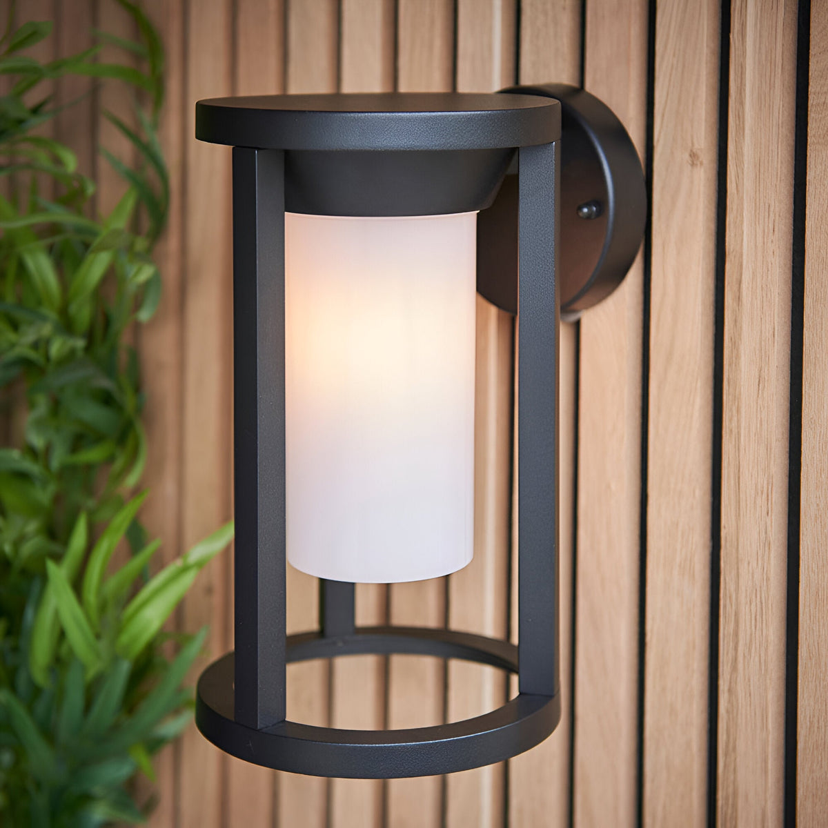 Endon Ashdale outdoor wall light with black aluminium frame and white polycarbonate cylinder shade, mounted on a slatted wood wall beside greenery.