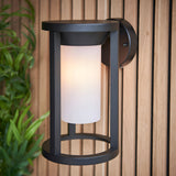 Endon Ashdale outdoor wall light with black aluminium frame and white polycarbonate cylinder shade, mounted on a slatted wood wall beside greenery.
