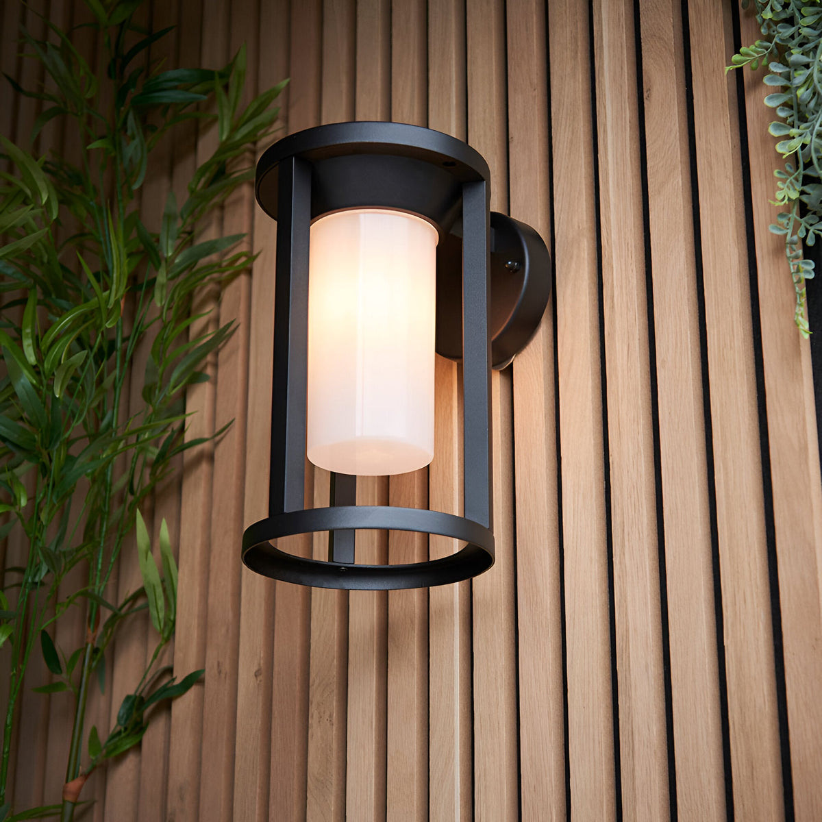 Endon Ashdale outdoor wall light with a cylindrical white polycarbonate shade in a matte black cage frame, mounted on a slatted wood wall beside foliage.