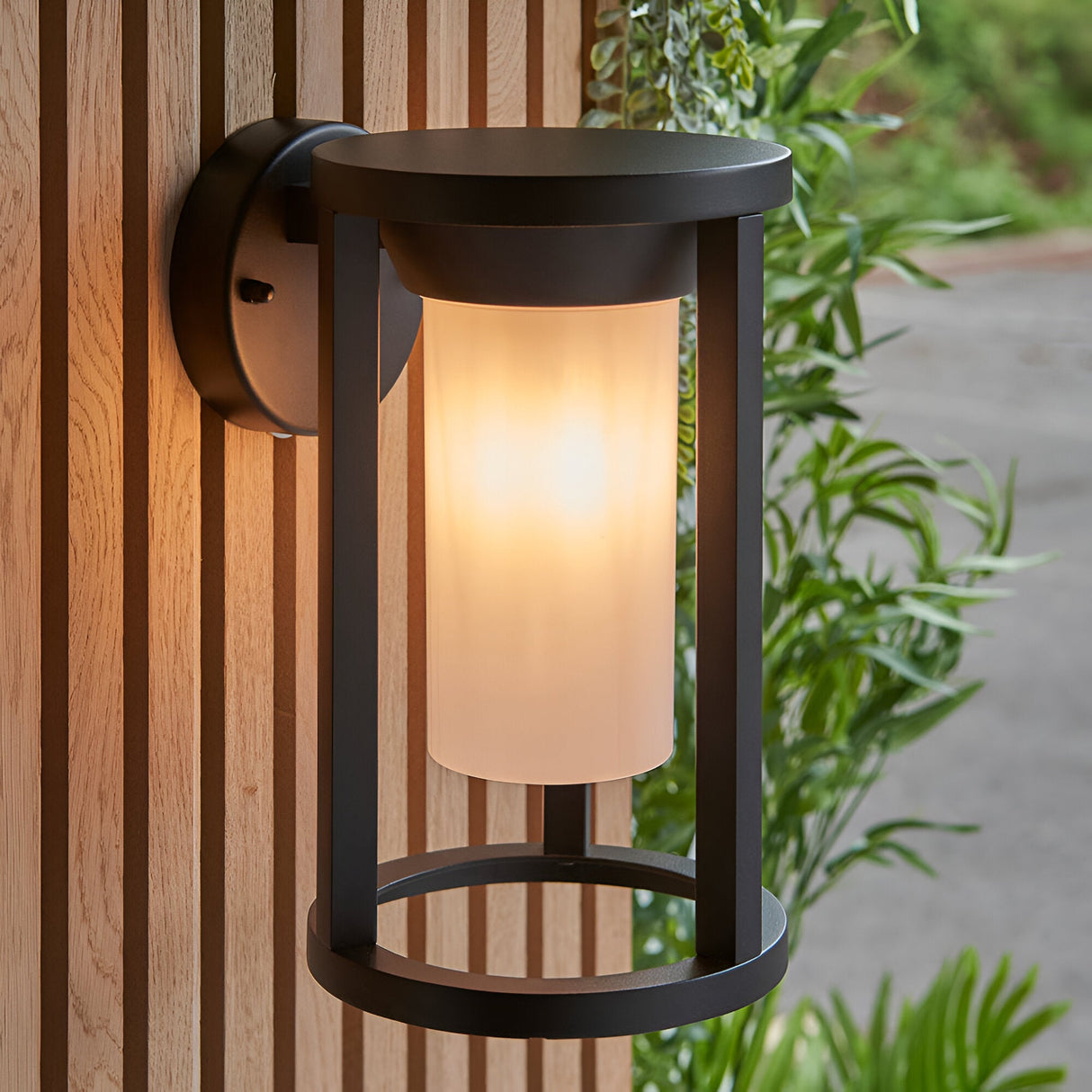 Endon Ashdale outdoor wall light in black aluminium with a cylindrical white polycarbonate shade, mounted on a slatted wood wall beside foliage.