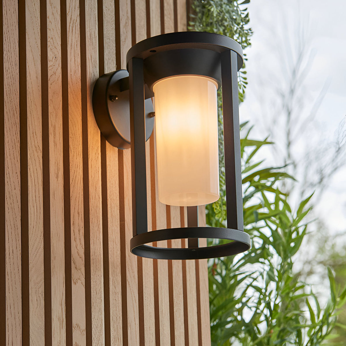 Endon Ashdale outdoor wall light with black aluminium frame and white polycarbonate shade, mounted on a slatted wood wall beside greenery.