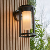 Endon Ashdale outdoor wall light with black aluminium frame and white polycarbonate shade, mounted on a slatted wood wall beside greenery.