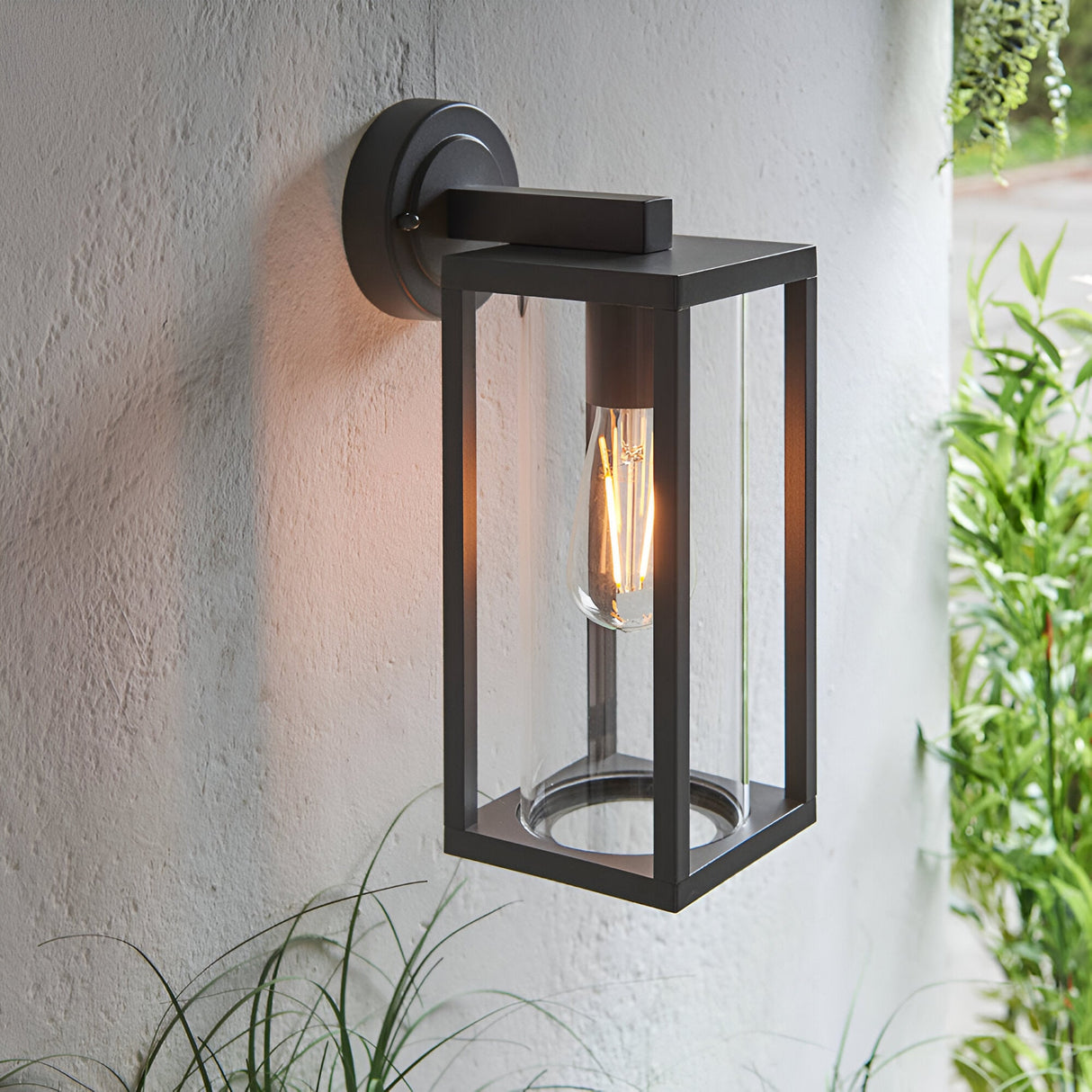 Endon Bramwood outdoor wall light in textured black aluminium with clear glass panels and a filament E27 bulb, mounted on a white exterior wall beside foliage.