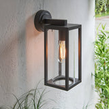 Endon Bramwood outdoor wall light in textured black aluminium with clear glass panels and a filament E27 bulb, mounted on a white exterior wall beside foliage.