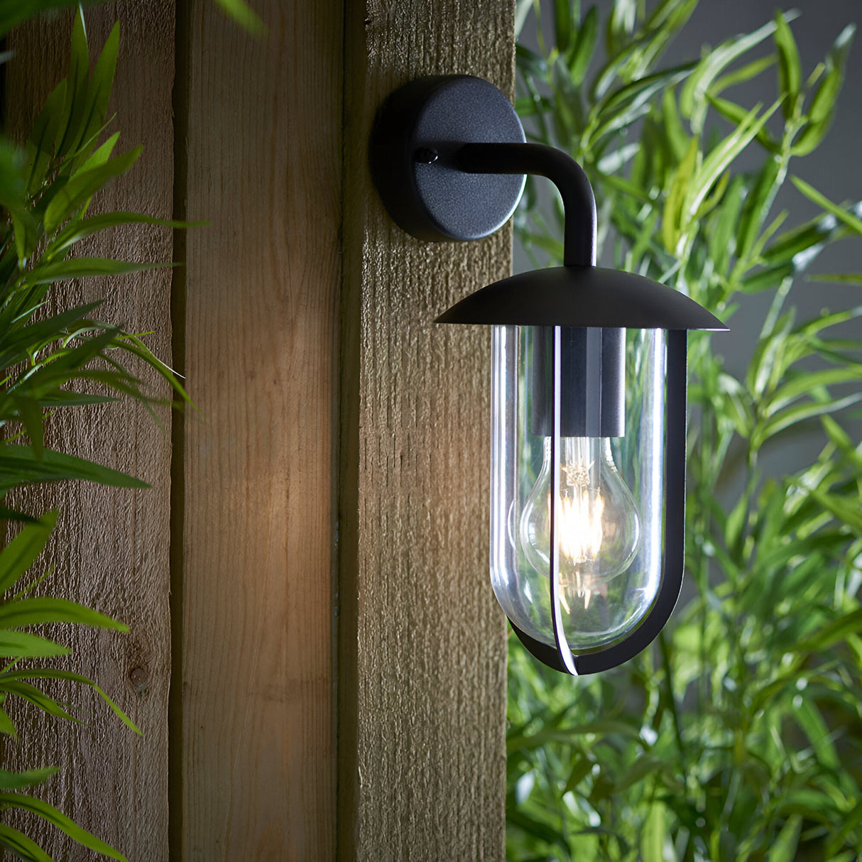 Chartley Outdoor Wall Light in black aluminium with curved arm, domed shade and clear glass shield, mounted on a wooden fence panel amid greenery.