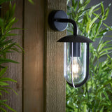 Chartley Outdoor Wall Light in black aluminium with curved arm, domed shade and clear glass shield, mounted on a wooden fence panel amid greenery.