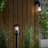 Black aluminium Chartley outdoor wall light with clear shade and filament bulb, mounted beside a matching bollard light and wooden panelling.