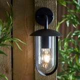 Chartley Outdoor Wall Light in black textured aluminium, mounted on wooden fence with a clear shade housing a lit E27 filament bulb, surrounded by greenery.