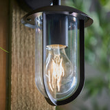 Chartley Outdoor Wall Light in black aluminium with a clear glass shade around an exposed E27 filament bulb, mounted on a rope-textured wall.