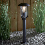 Chartley Outdoor Post Light in black textured aluminium with a domed top and clear polycarbonate shade housing a lit bulb, set on gravel amid tall grasses.