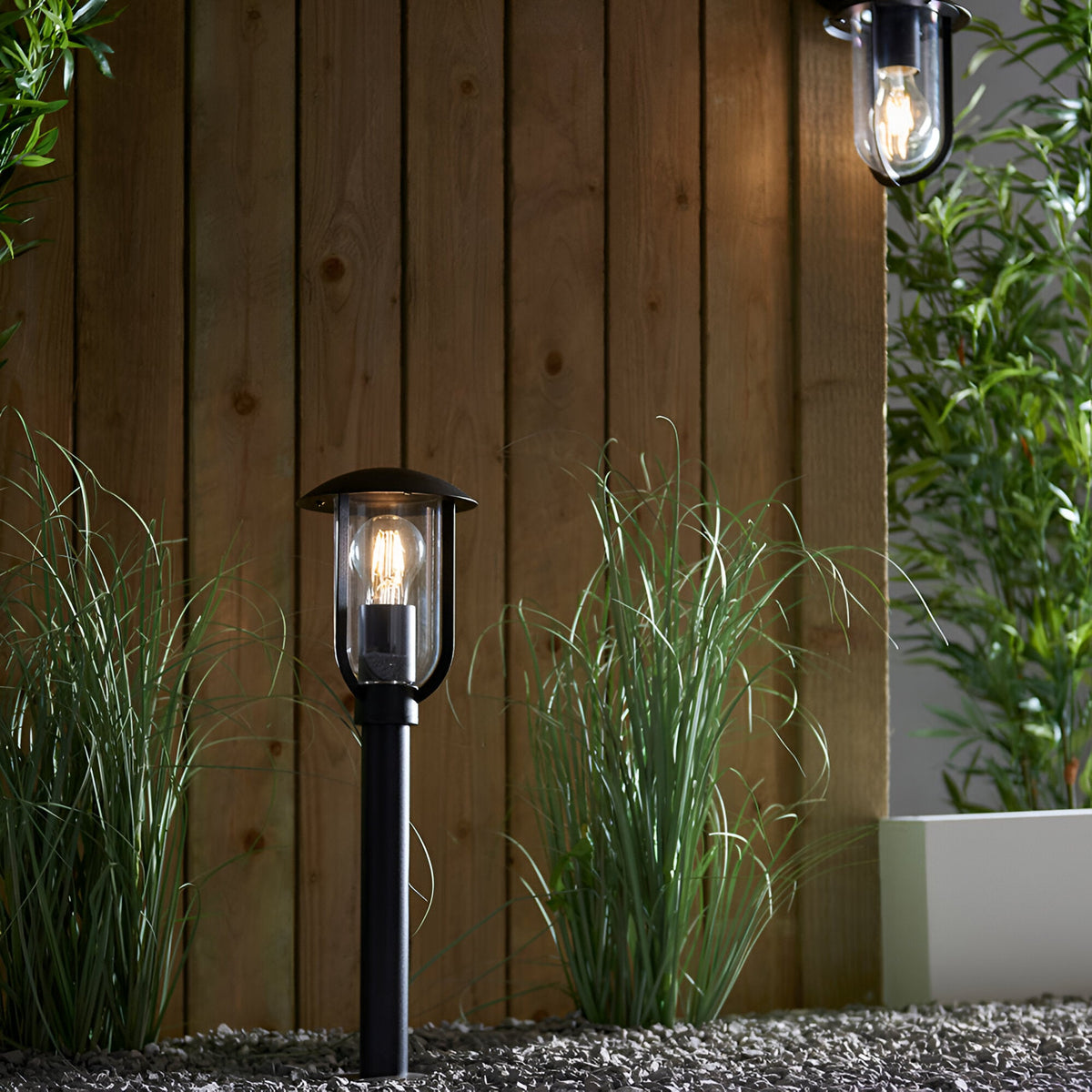 Chartley Outdoor Post Light in black aluminium with clear polycarbonate shade, lit and set among garden grasses against a wooden fence, IP44 rated.