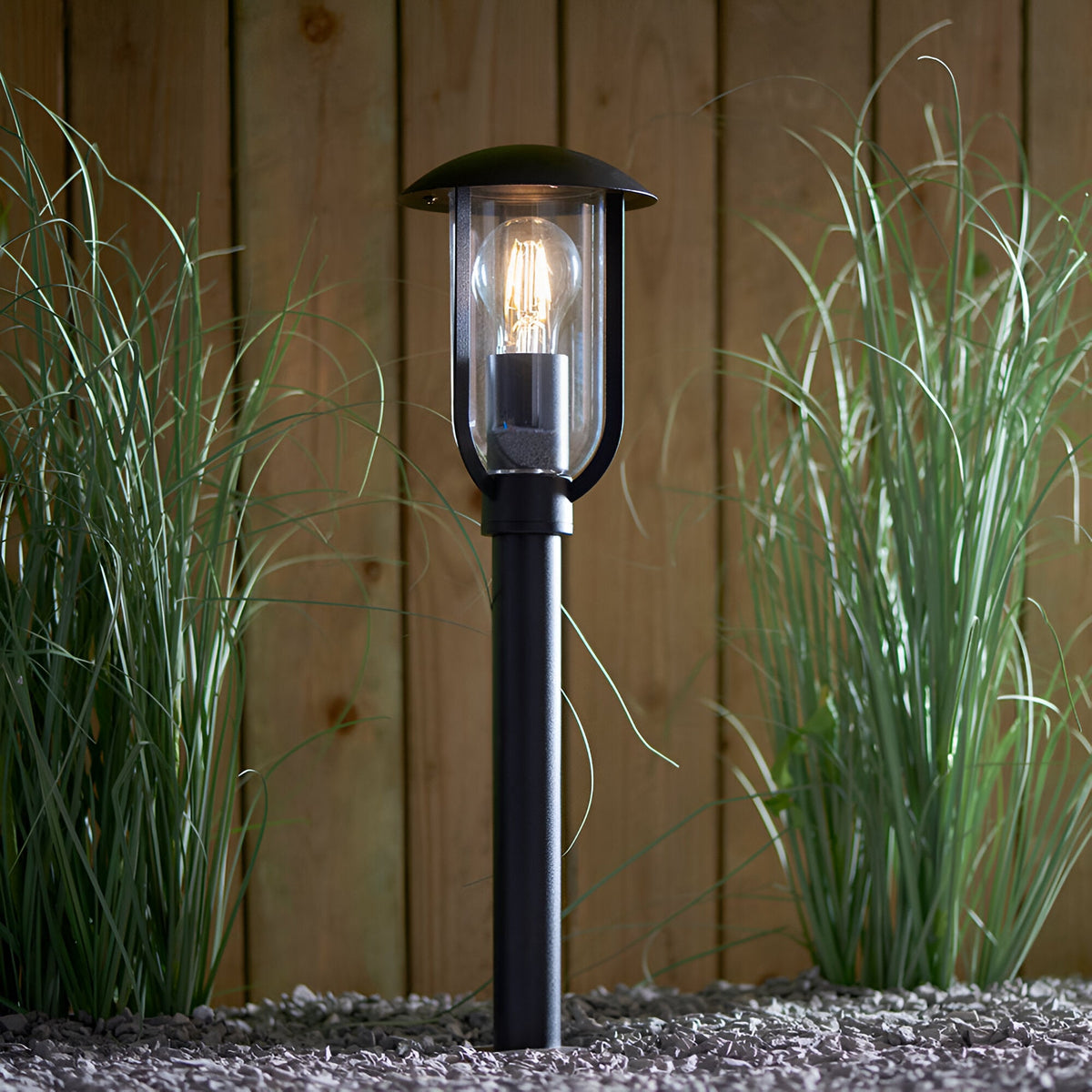 Chartley Outdoor Post Light in black aluminium with clear polycarbonate shade and lit E27 bulb, set in gravel among grasses against a wooden fence.