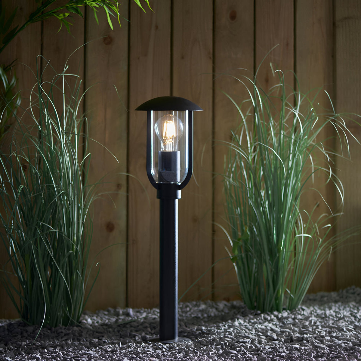 Chartley Outdoor Post Light in black textured aluminium with clear polycarbonate shade and lit E27 bulb, set among garden grasses against a wooden fence.